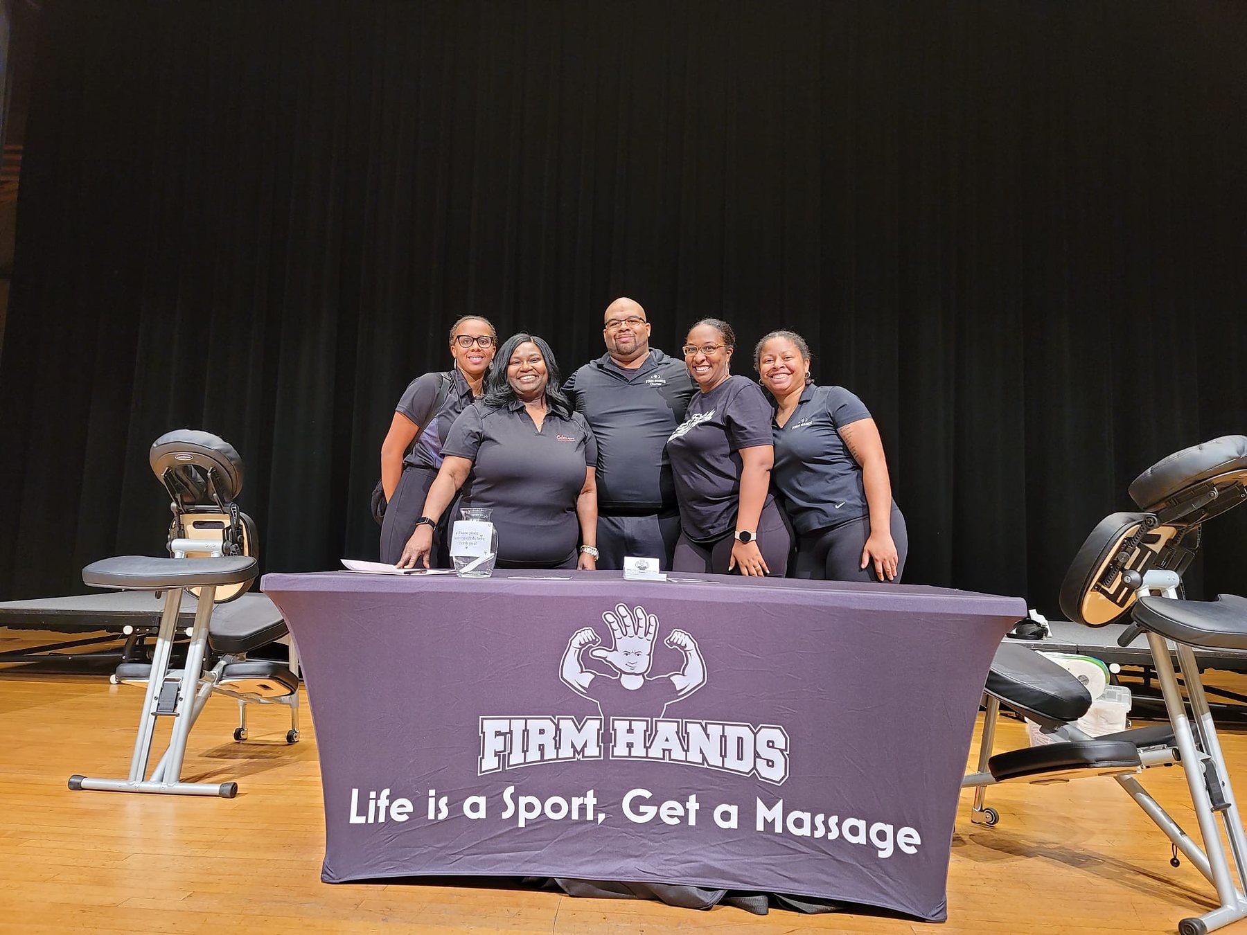 Five Firm Hands therapists behind their table on an auditorium stage, massage chairs either side