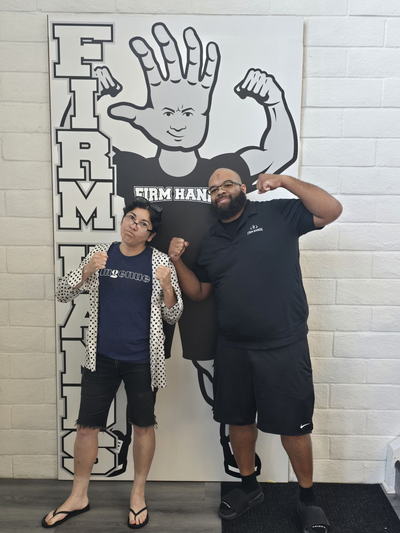 Two people flexing beside the life-size Bruce board, one in a Firm Hands shirt