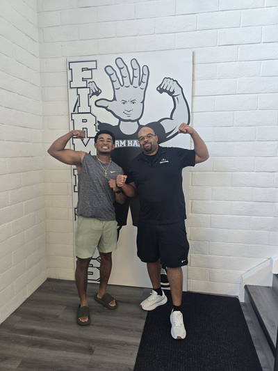 Two people flexing beside the Bruce board, one in a Firm Hands shirt